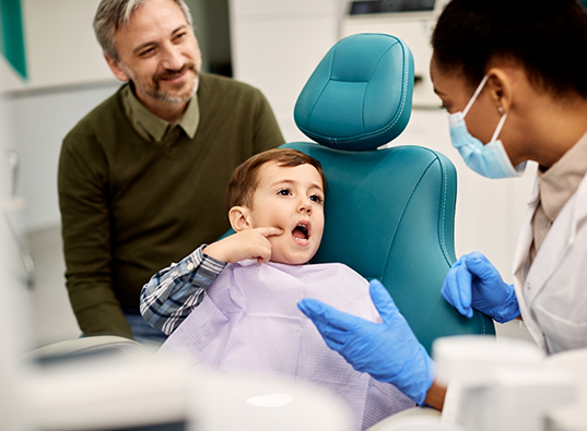 Young boy in dental chair pointing to the side of his face while talking to dentist