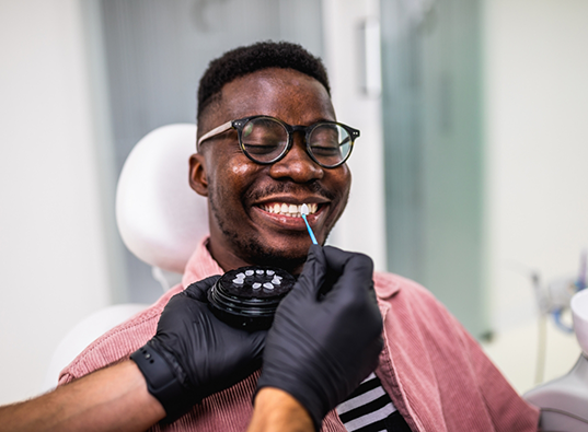 Man in dental chair smiling while dentist holds a veneer up to his teeth