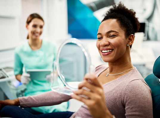 Woman in dental chair holding a mirror and looking at her smile