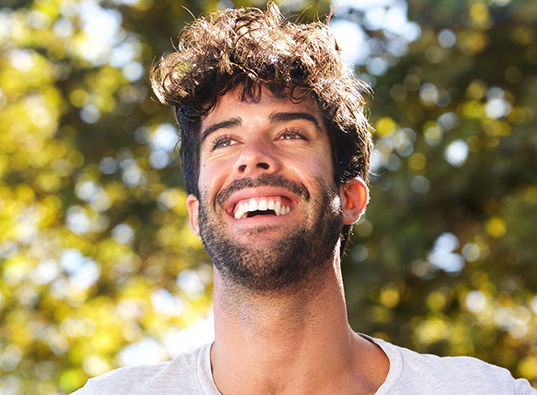 Young man with curly dark hair smiling in a forest on a sunny day